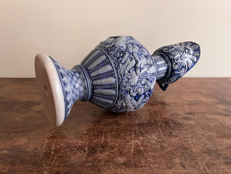 May include: A blue and white ceramic vase with intricate relief detailing. The vase features a wide base, a central body adorned with cherubic figures, and a spout with ornate patterns. The color scheme is a classic blue and white.