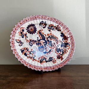 Könnte beinhalten: Eine ovale Keramikplatte mit gewelltem Rand auf weißem Grund, verziert mit komplexen Blumen- und Füllhornmustern in tiefem Marineblau und gebranntem Orange. Der Rand ist mit einem wiederkehrenden Schnörkelmotiv in passenden Farben versehen.