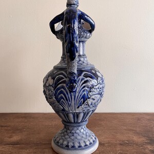 May include: A blue and white ceramic pitcher with a detailed relief design. The pitcher features a handle with a sculpted figure and ornate patterns. The base has a textured, scaled pattern. The overall design is classical and decorative.