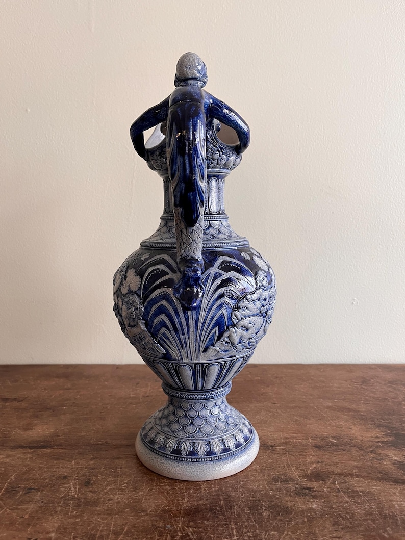 May include: A blue and white ceramic pitcher with a detailed relief design. The pitcher features a handle with a sculpted figure and ornate patterns. The base has a textured, scaled pattern. The overall design is classical and decorative.