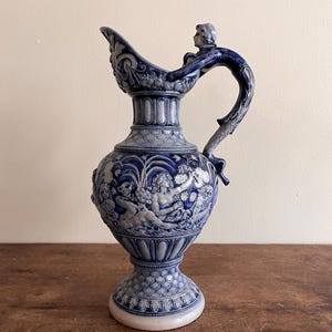 May include: A blue and white ceramic pitcher with a detailed relief design. The pitcher features a handle with a sculpted figure, a wide spout, and a bulbous body adorned with classical figures. The base is a pedestal design.