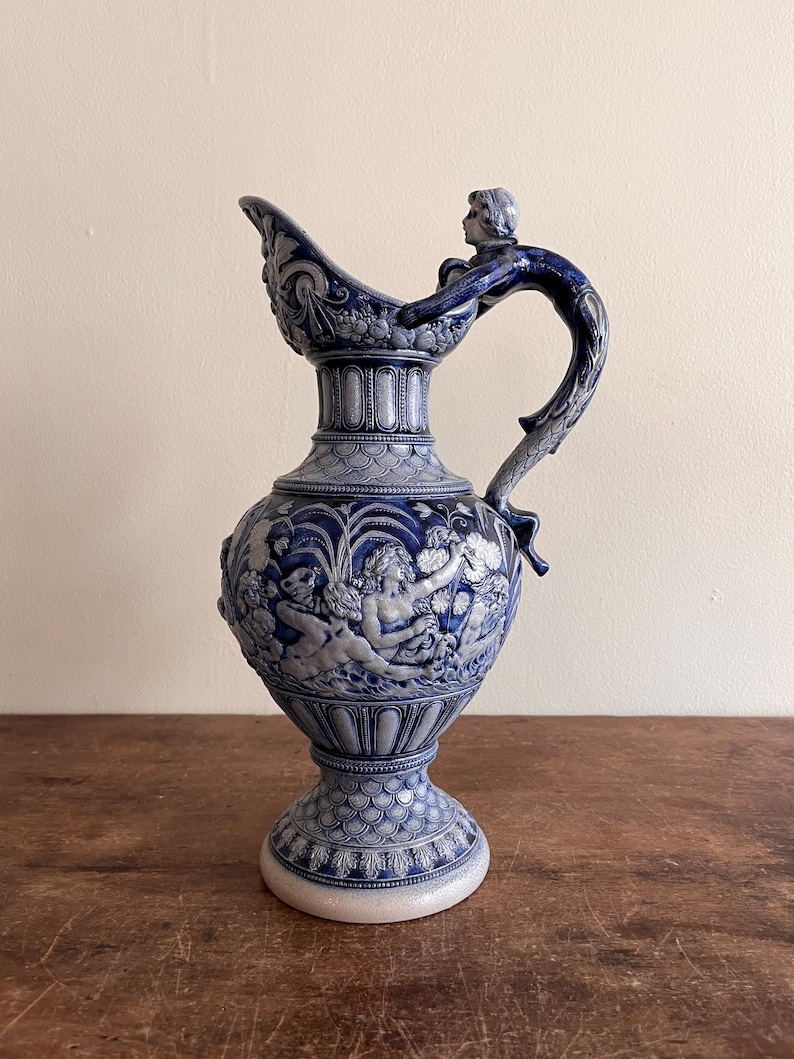 May include: A blue and white ceramic pitcher with a detailed relief design. The pitcher features a handle with a sculpted figure, a wide spout, and a bulbous body adorned with classical figures. The base is a pedestal design.