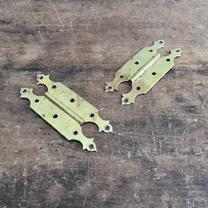 May include: Two antique brass hinges with ornate designs. The hinges are on a wooden surface.
