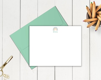 May include: A white stationery set featuring a rainbow design and the name "Olivia" in a light blue font. The set includes a matching light green envelope, a pair of gold scissors, and a small container of colored pencils.