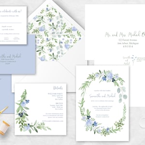 Puede incluir: Conjunto de papelería de boda con diseños florales en acuarela. Incluye invitaciones, tarjetas RSVP, sobres y tarjetas de detalles. El diseño incorpora flores azules y follaje verde, con tijeras doradas y un carrete de hilo.