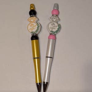 May include: Two pens with decorative ring toppers. One pen is gold with a black ring topper and the other is white with a pink ring topper. Both ring toppers have a diamond design and the text "She Said Yes!"