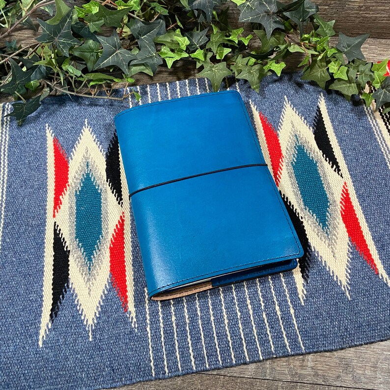 May include: A bright blue leather notebook with an elastic closure. The notebook is lying on a blue and white patterned rug.