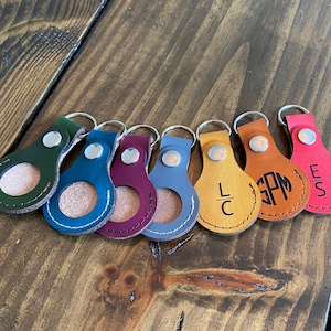 May include: Seven leather keychains with a circular cutout in the center. Each keychain has a different color and a monogrammed initial in the center. The keychains are arranged in a row on a wooden surface.