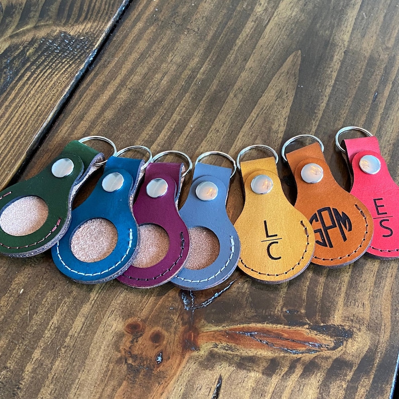 May include: Seven leather keychains with a circular cutout in the center. Each keychain has a different color and a monogrammed initial in the center. The keychains are arranged in a row on a wooden surface.