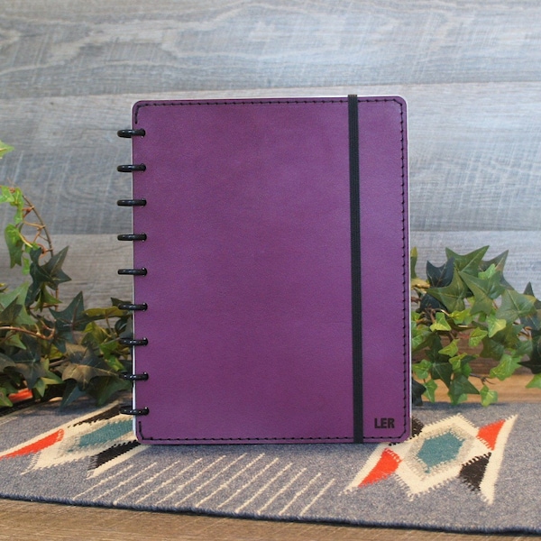May include: A purple leather-bound notebook with a black elastic closure and a black coil binding. The notebook has the word "LER" embossed on the bottom right corner.