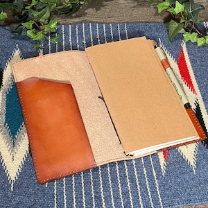 May include: A brown leather notebook cover with a pen holder and a pocket on the front. The cover is open to reveal a blank notebook.