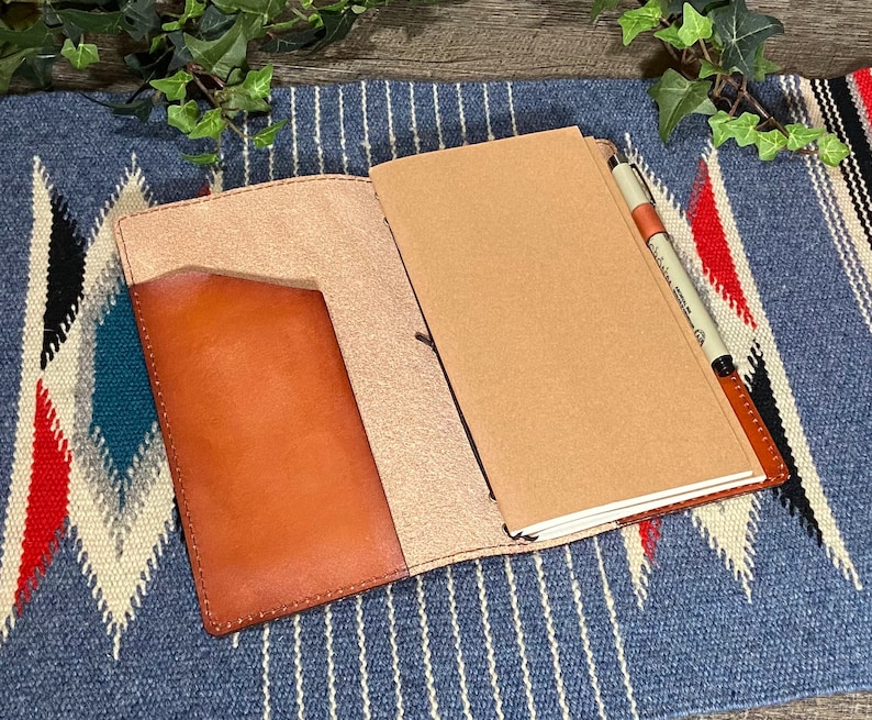 May include: A brown leather notebook cover with a pen holder and a pocket on the front. The cover is open to reveal a blank notebook.