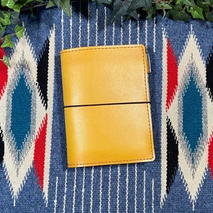 May include: A yellow leather bound journal with a brown elastic closure. The journal is sitting on a blue and white patterned surface.