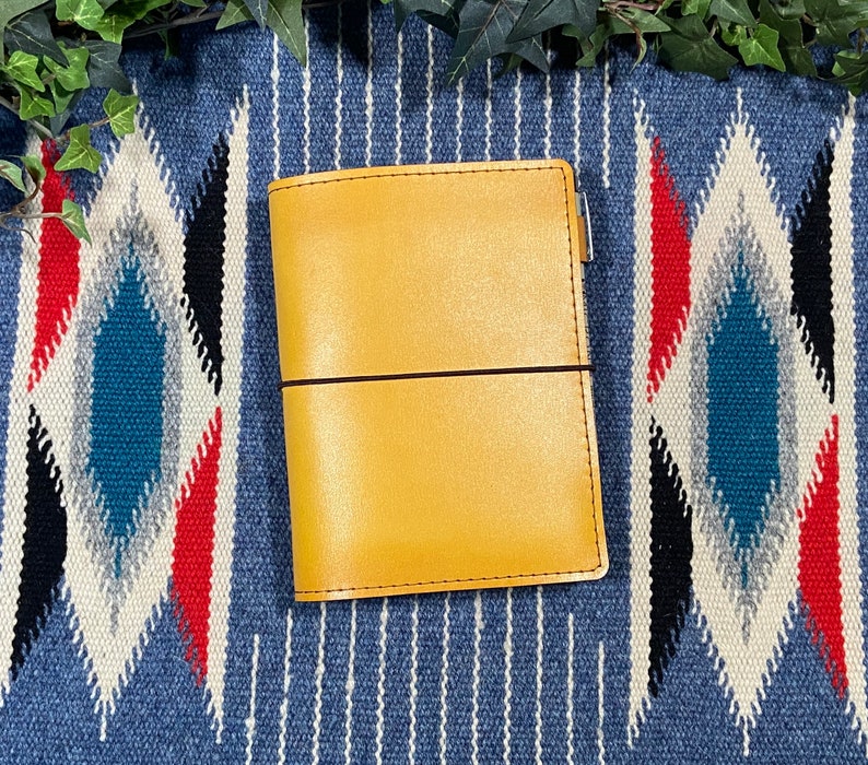 May include: A yellow leather bound journal with a brown elastic closure. The journal is sitting on a blue and white patterned surface.