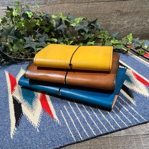 May include: Three leather-bound journals stacked on top of each other. The top journal is yellow, the middle journal is brown, and the bottom journal is blue.