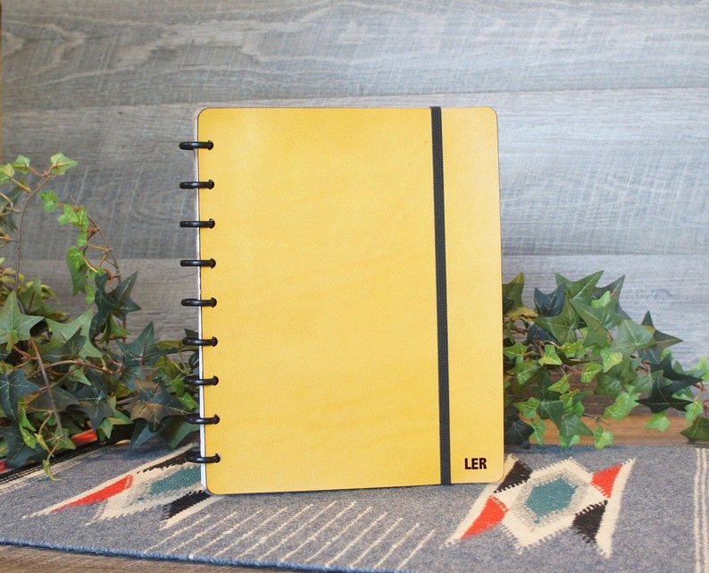 May include: A yellow leather bound notebook with a black elastic closure and a black coil binding. The notebook has the word "LER" printed on the bottom right corner.