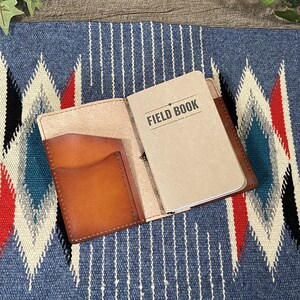 May include: A brown leather notebook cover with a pocket on the front. The cover is open to reveal a blank notebook with the text "FIELD BOOK" printed on the front.