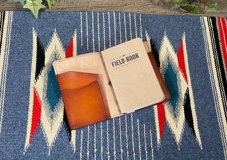 May include: A brown leather notebook cover with a pocket on the front. The cover is open to reveal a blank notebook with the text "FIELD BOOK" printed on the front.