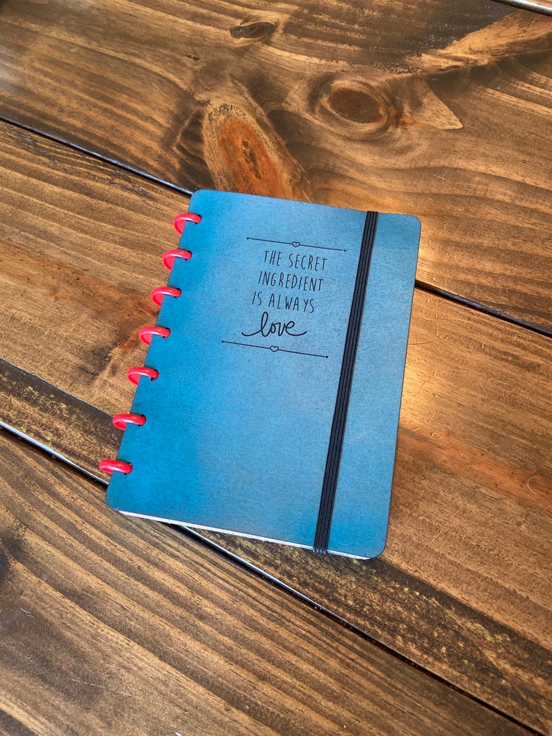 May include: A blue notebook with a black elastic band closure. The notebook has a quote that reads "The secret ingredient is always love" in black lettering.