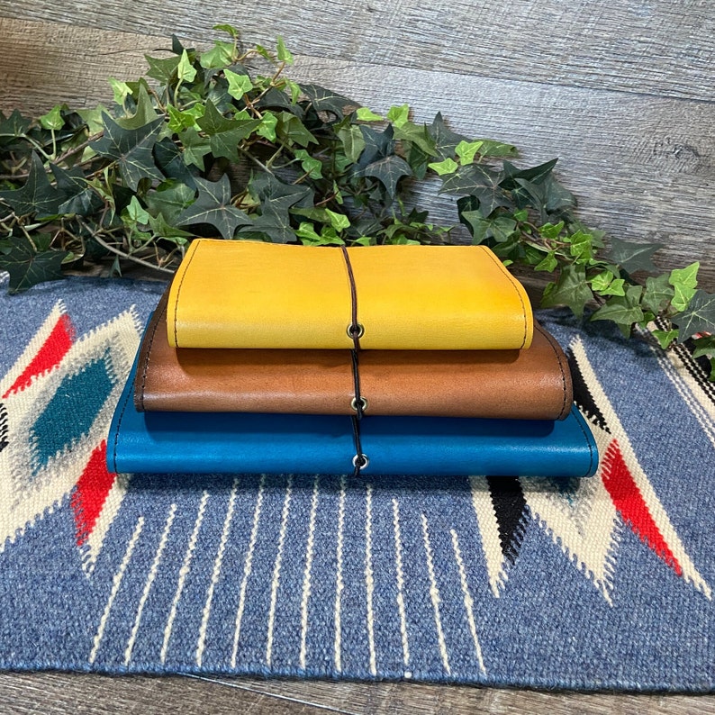 May include: Three leather-bound journals stacked on top of each other. The top journal is yellow, the middle journal is brown, and the bottom journal is blue.