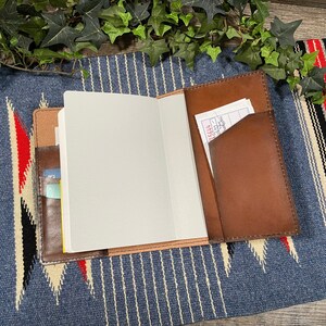 May include: A brown leather journal with a blank white page open. The journal has a pocket on the outside for holding cards or receipts. The journal is lying on a blue and white striped fabric.