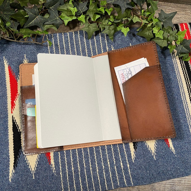 May include: A brown leather journal with a blank white page open. The journal has a pocket on the outside for holding cards or receipts. The journal is lying on a blue and white striped fabric.