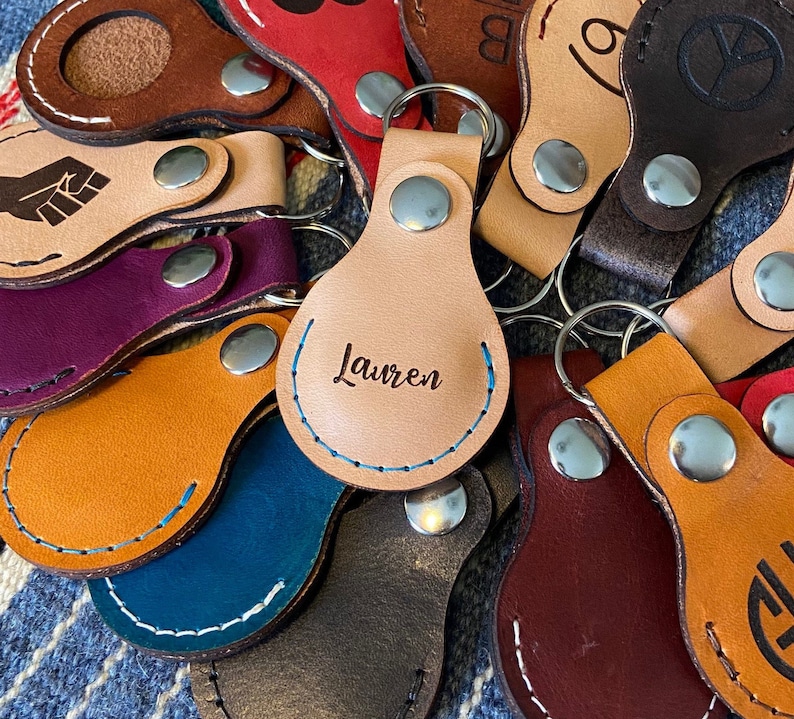 May include: A collection of leather keychains with various designs, including a fist, a peace sign, and a name "Lauren".