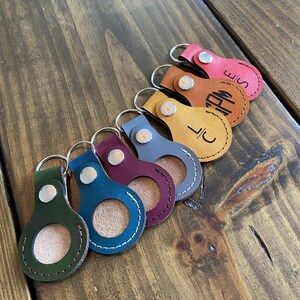 May include: Seven leather keychains with a circular cutout in the center. The keychains are in various colors, including green, blue, burgundy, gray, tan, orange, and red. Each keychain has a silver ring and a silver snap closure. The keychains are arranged in a row on a wooden surface.