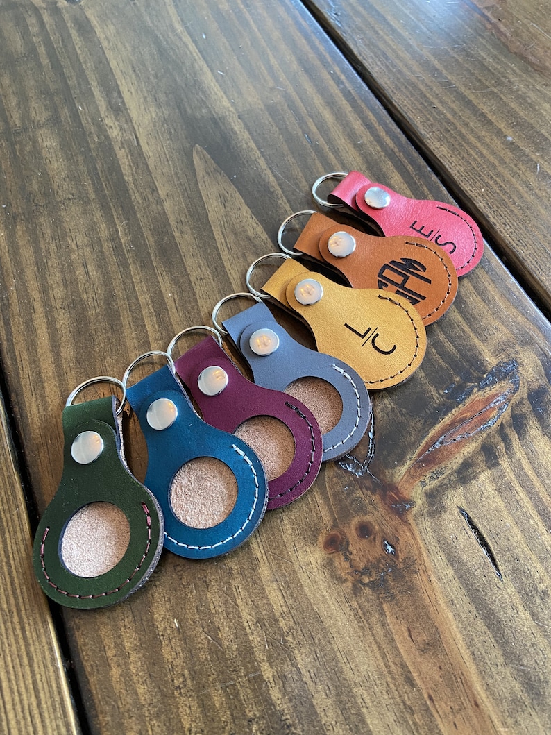 May include: Seven leather keychains with a circular cutout in the center. The keychains are in various colors, including green, blue, burgundy, gray, tan, orange, and red. Each keychain has a silver ring and a silver snap closure. The keychains are arranged in a row on a wooden surface.