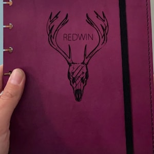 Op de afbeelding: Een diep paars lederen dagboek met een zwarte elastische sluiting. De voorkant heeft een gegraveerd ontwerp van een hertenschedel met geweien en het woord "REDWIN" erboven.
