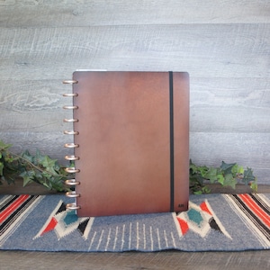 May include: A brown leather bound journal with a black elastic closure and silver rings. The journal is sitting on a blue and red patterned rug.