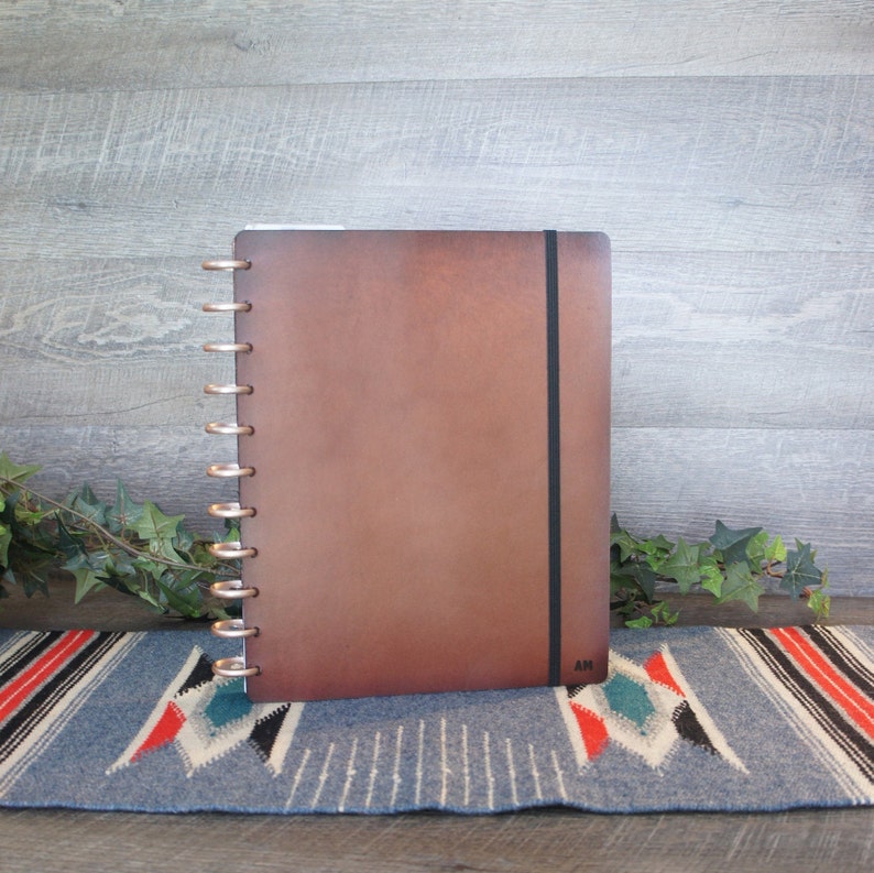 May include: A brown leather bound journal with a black elastic closure and silver rings. The journal is sitting on a blue and red patterned rug.