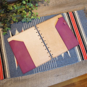 May include: A tan leather bound journal with a purple leather flap on the front and back covers. The journal is open and lying on a blue and red striped fabric.