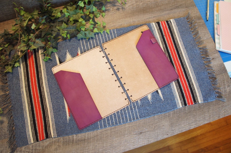 May include: A tan leather bound journal with a purple leather flap on the front and back covers. The journal is open and lying on a blue and red striped fabric.