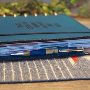 May include: A teal leather bound planner with a black elastic band and a silver pen. The planner is filled with white paper and has a variety of colorful tabs.