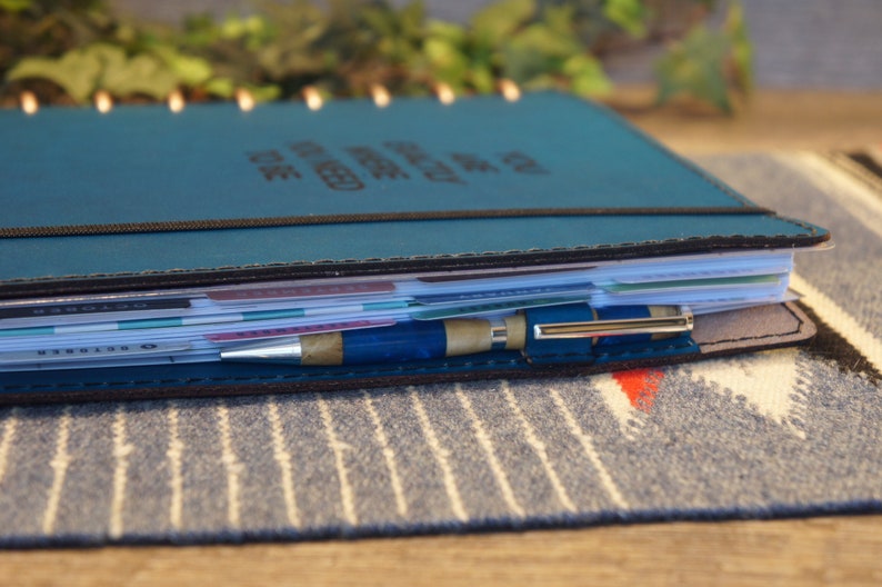 May include: A teal leather bound planner with a black elastic band and a silver pen. The planner is filled with white paper and has a variety of colorful tabs.