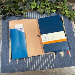 May include: A blue leather notebook cover with a brown leather flap and a pocket for cards or cash. The notebook cover is open to reveal a blue Moleskine notebook with the text "Classic Collection" and "Ruled Notebook carnet 19x13 cm".