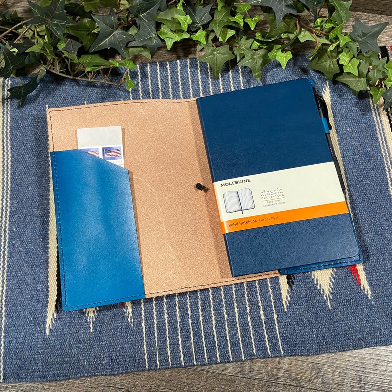 May include: A blue leather notebook cover with a brown leather flap and a pocket for cards or cash. The notebook cover is open to reveal a blue Moleskine notebook with the text "Classic Collection" and "Ruled Notebook carnet 19x13 cm".