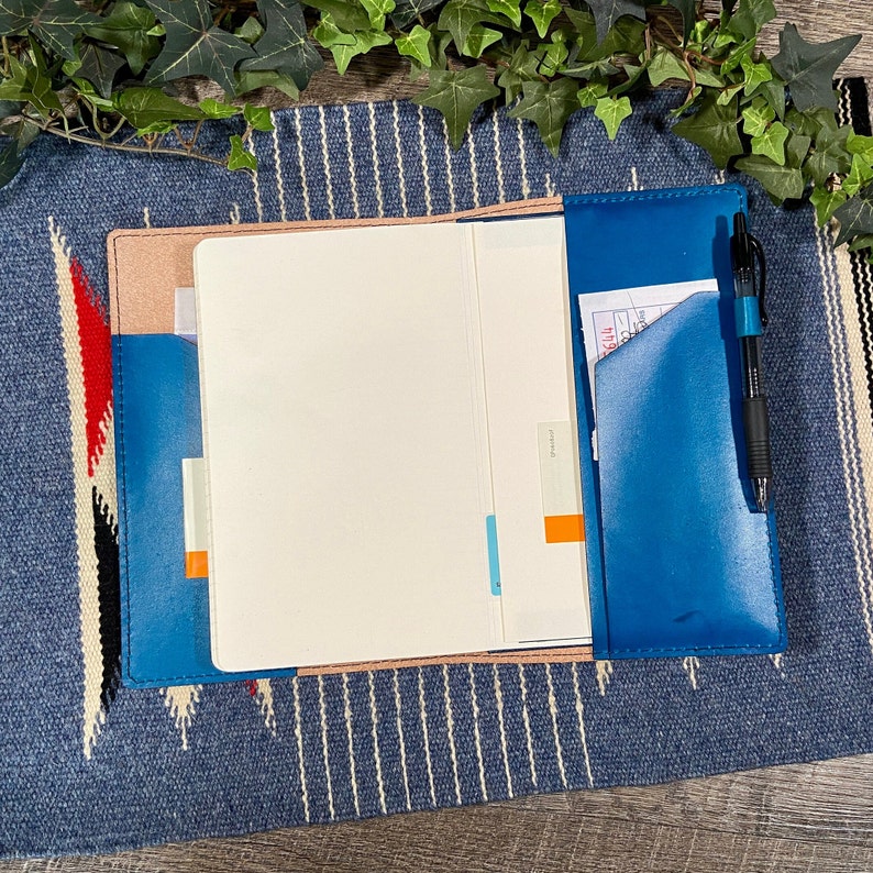 May include: A blue leather notebook with a pink interior and a pen in the pocket. The notebook is open and has blank pages.