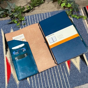 May include: A teal leather notebook cover with a pocket for cards and a separate pocket for a phone. The cover is open to reveal a tan leather interior and a blue Moleskine notebook with the text "Classic Collection" and "Pocket Notebook" printed on the cover.