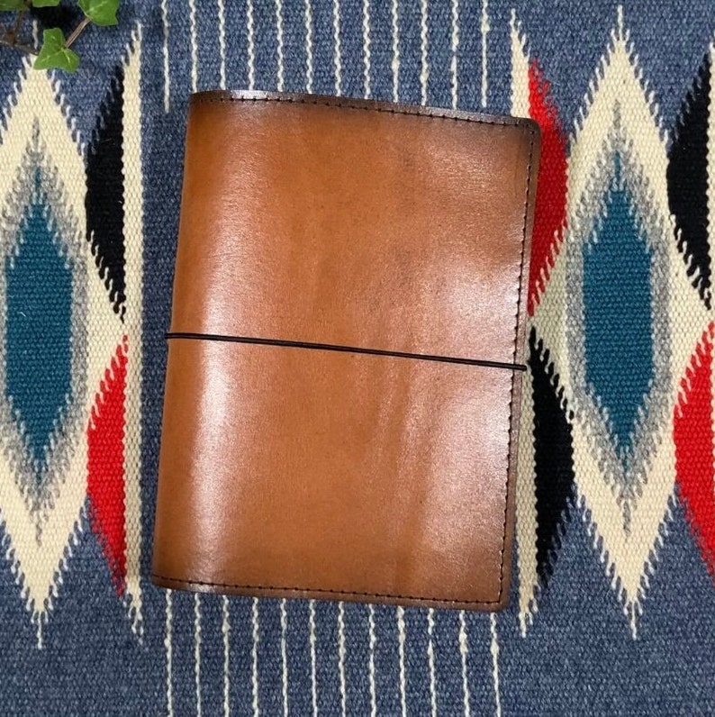 May include: A brown leather notebook cover with a black elastic band closure. The cover is on a patterned fabric background.