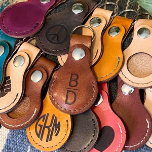 May include: A collection of leather keychains with various designs and engravings. Some keychains feature a round shape with a cutout for a coin, while others have a more traditional keychain shape. The keychains are made of brown, tan, and cream leather and feature engravings such as "B/D", "GKM", and a peace sign.