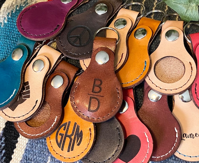 May include: A collection of leather keychains with various designs and engravings. Some keychains feature a round shape with a cutout for a coin, while others have a more traditional keychain shape. The keychains are made of brown, tan, and cream leather and feature engravings such as "B/D", "GKM", and a peace sign.