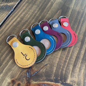 May include: Six leather keychains in different colors, including yellow, green, blue, burgundy, gray, and red. Each keychain has a silver ring and a small circle cut out of the leather. The yellow keychain has the initials "LC" engraved on it.