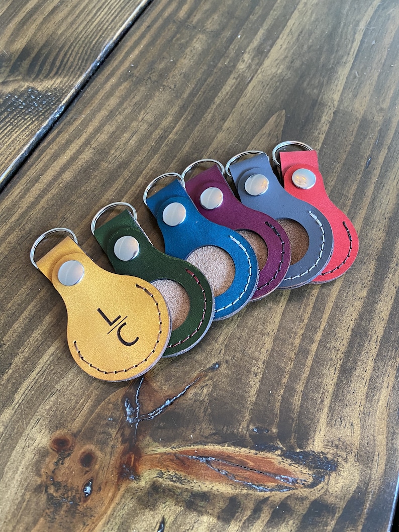 May include: Six leather keychains in different colors, including yellow, green, blue, burgundy, gray, and red. Each keychain has a silver ring and a small circle cut out of the leather. The yellow keychain has the initials "LC" engraved on it.