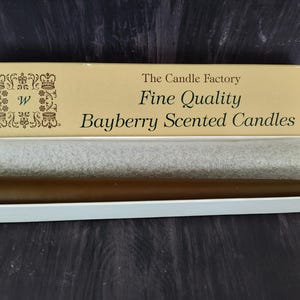 Puede incluir: Una vela marrón con aroma a bayberry en una caja blanca. La caja tiene el texto "The Candle Factory Fine Quality Bayberry Scented Candles". La vela está envuelta en papel de seda gris.