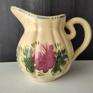 Puede incluir: Jarra de cerámica color crema con diseño floral. La jarra presenta una gran rosa burdeos y rosa con hojas verdes. El borde está decorado con puntos azules. El asa es curva.