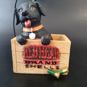 May include: A black Labrador Retriever figurine peeks out of a wooden crate. The crate has the words "REDBONE BRAND SHELLS" in red lettering. Two green and gold shotgun shells are in front of the crate. The dog wears an orange collar.