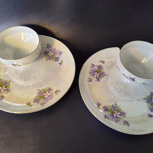 Pode incluir: Duas chávenas de chá e pires a condizer. As chávenas e os pratos de porcelana branca apresentam um delicado padrão floral em tons de roxo e verde. As bordas são adornadas com ouro. Os pratos têm uma forma única, projetada para segurar a chávena.