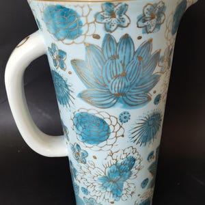 May include: A white ceramic pitcher featuring a repeating pattern of blue floral motifs and lotus blossoms. The design is accented with fine gold outlines throughout the surface. The pitcher has a curved handle and a wide pouring spout at the top.